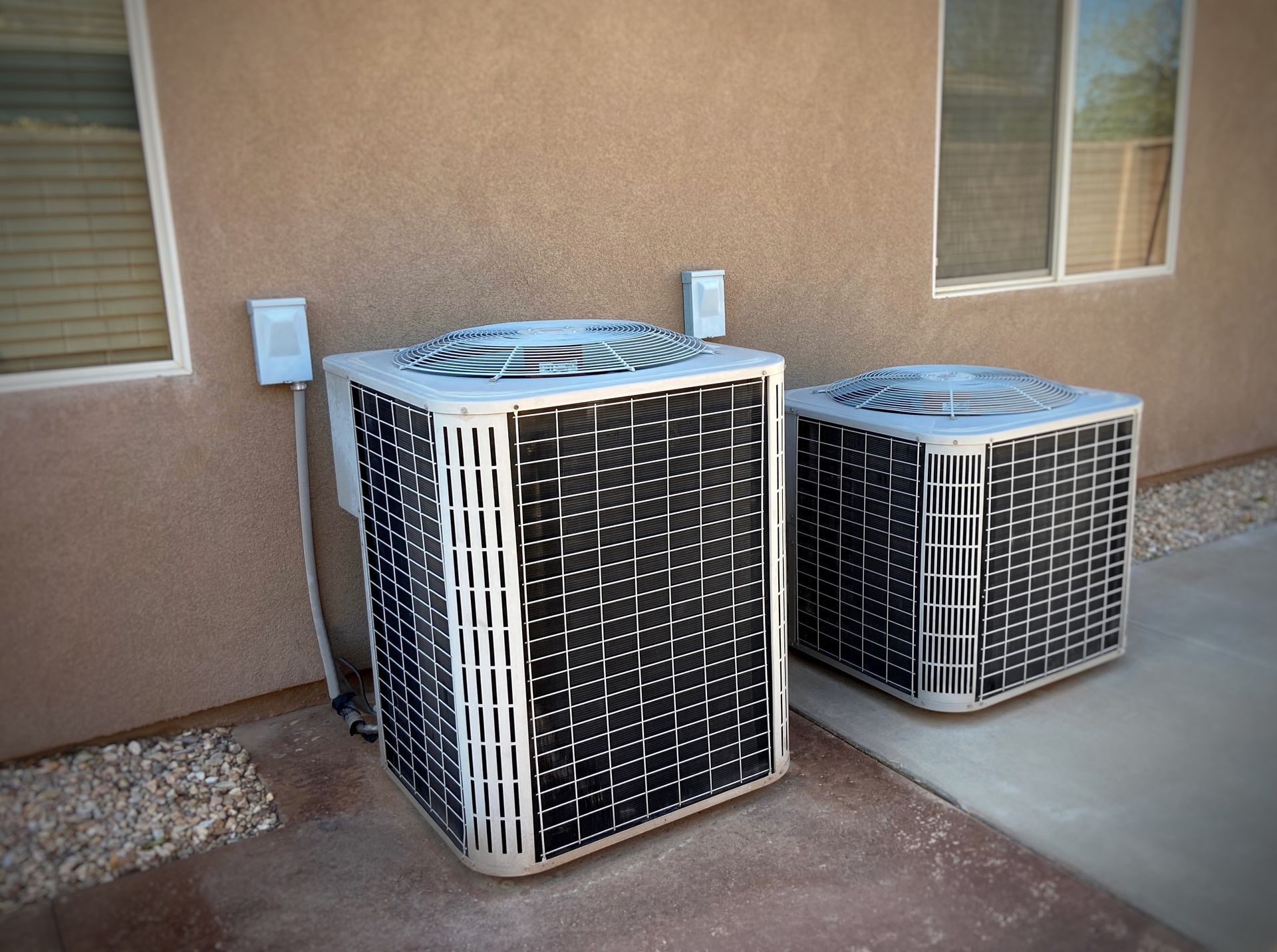 Two air conditioning units near a stucco wall and two windows.