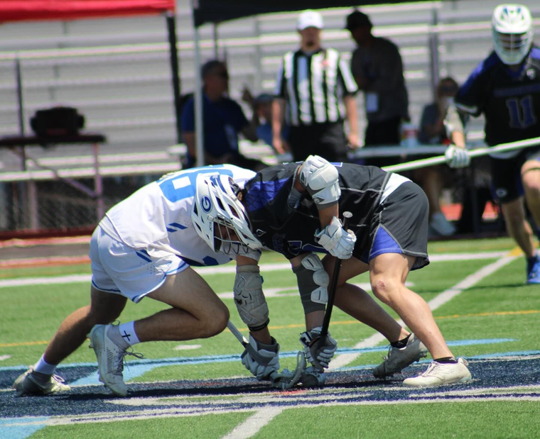 Two lacrosse players in white and dark uniforms crouch at the faceoff on a field.