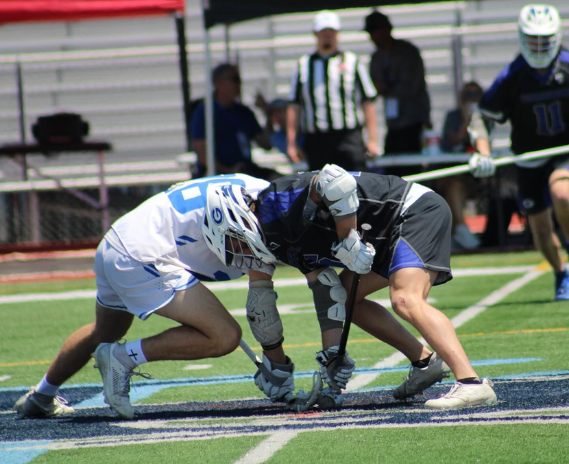 Two lacrosse players in white and dark uniforms crouch at the faceoff on a field.