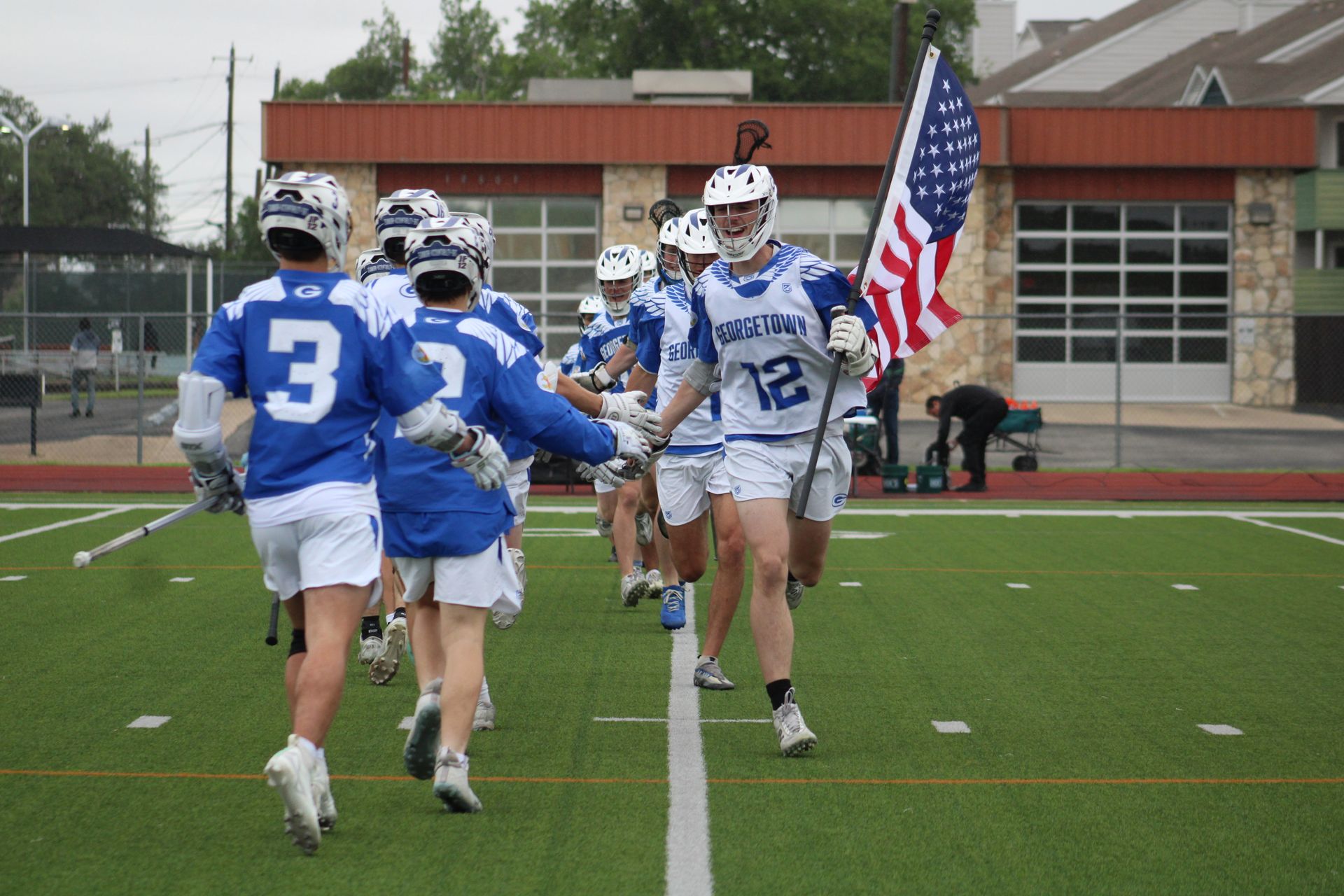 Lacrosse players in blue and white uniforms run on a field toward the camera, one carrying an American flag.