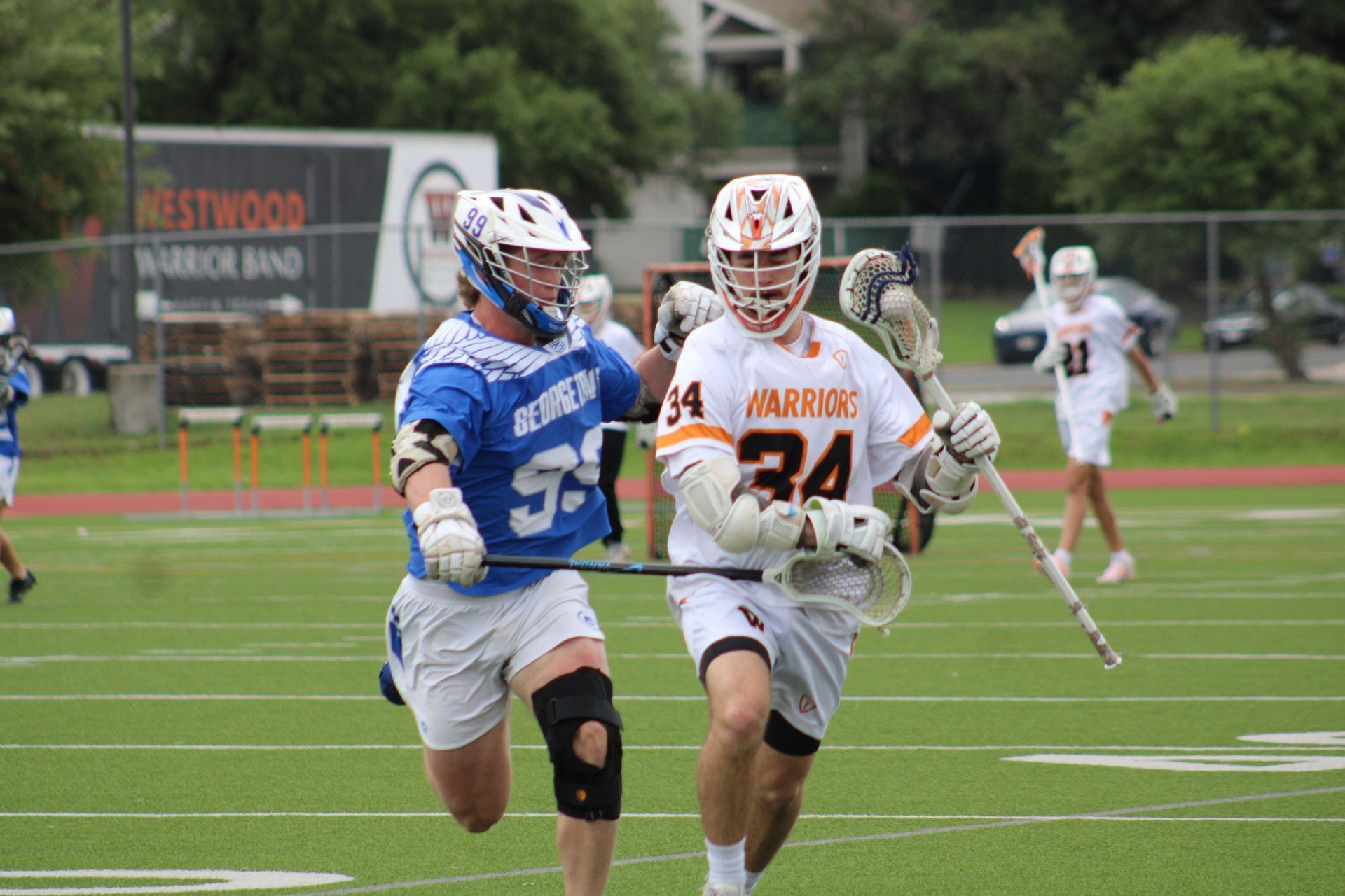 Two lacrosse players sprint on a field, one in blue and one in white and orange, chasing the ball.
