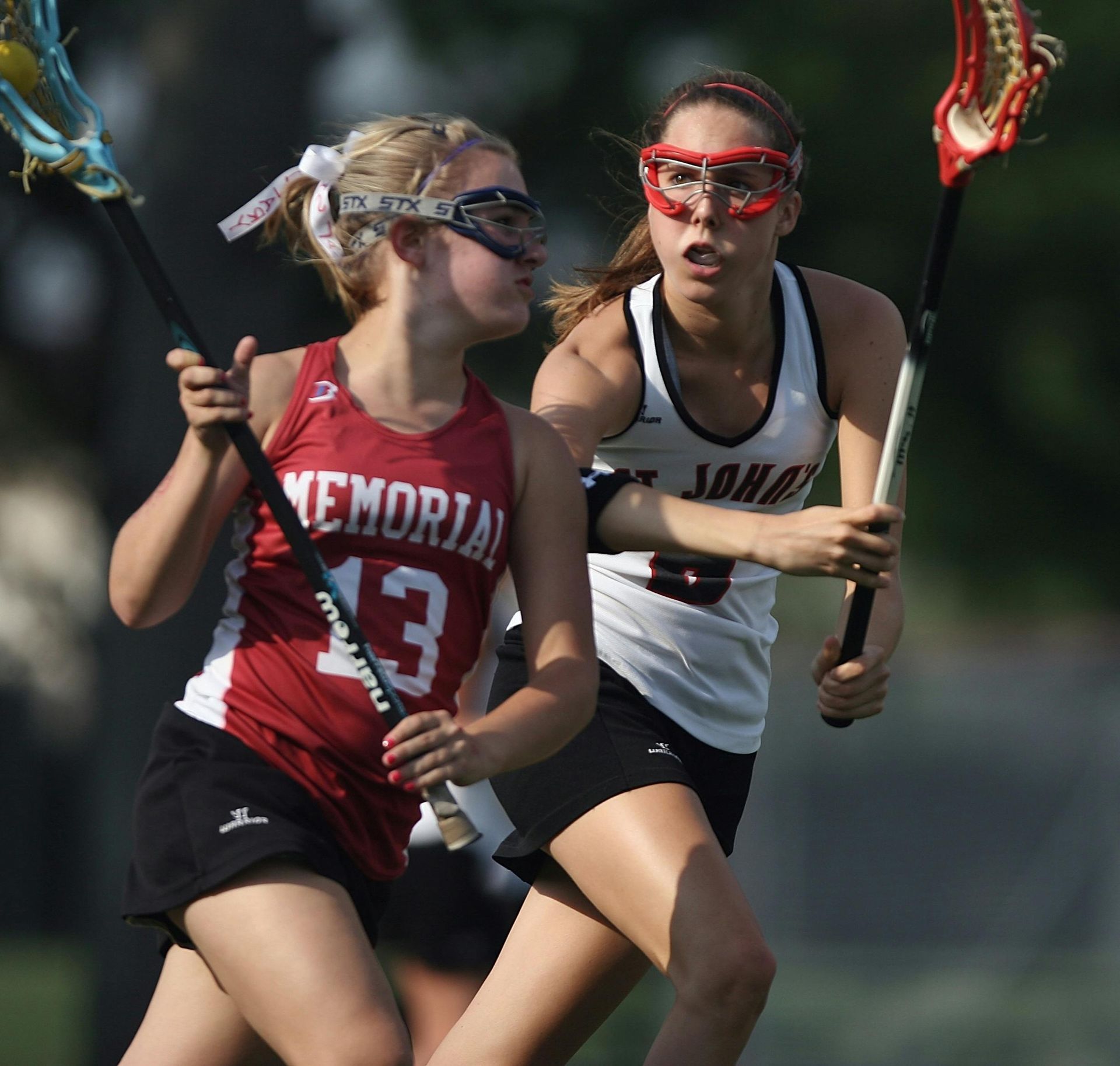Two lacrosse players compete, one in red and one in white, both holding sticks and wearing protective goggles.