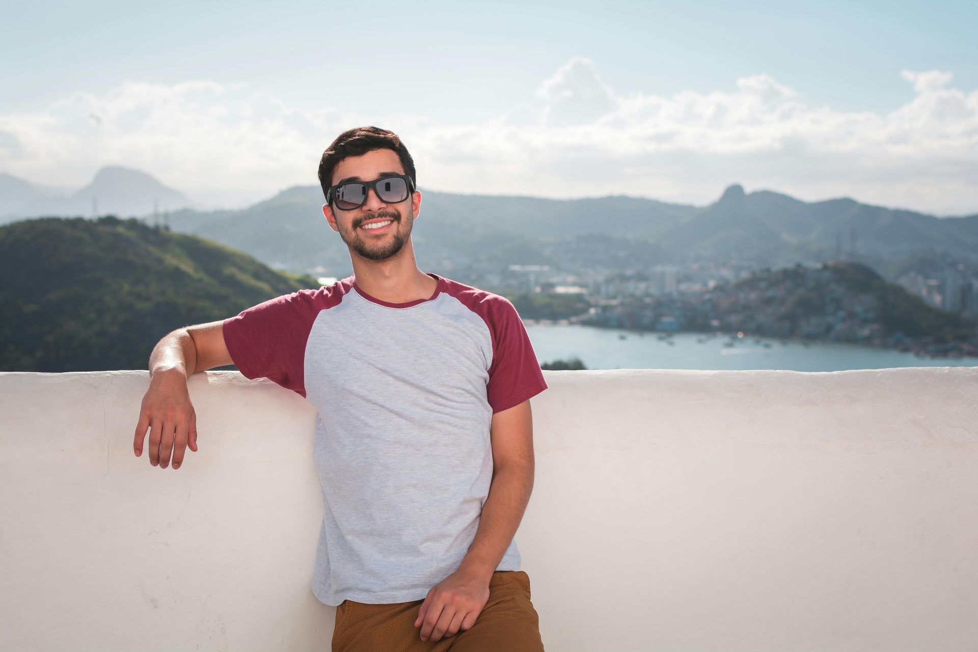 Man in sunglasses smiles, leans on wall; mountains and city visible in background.