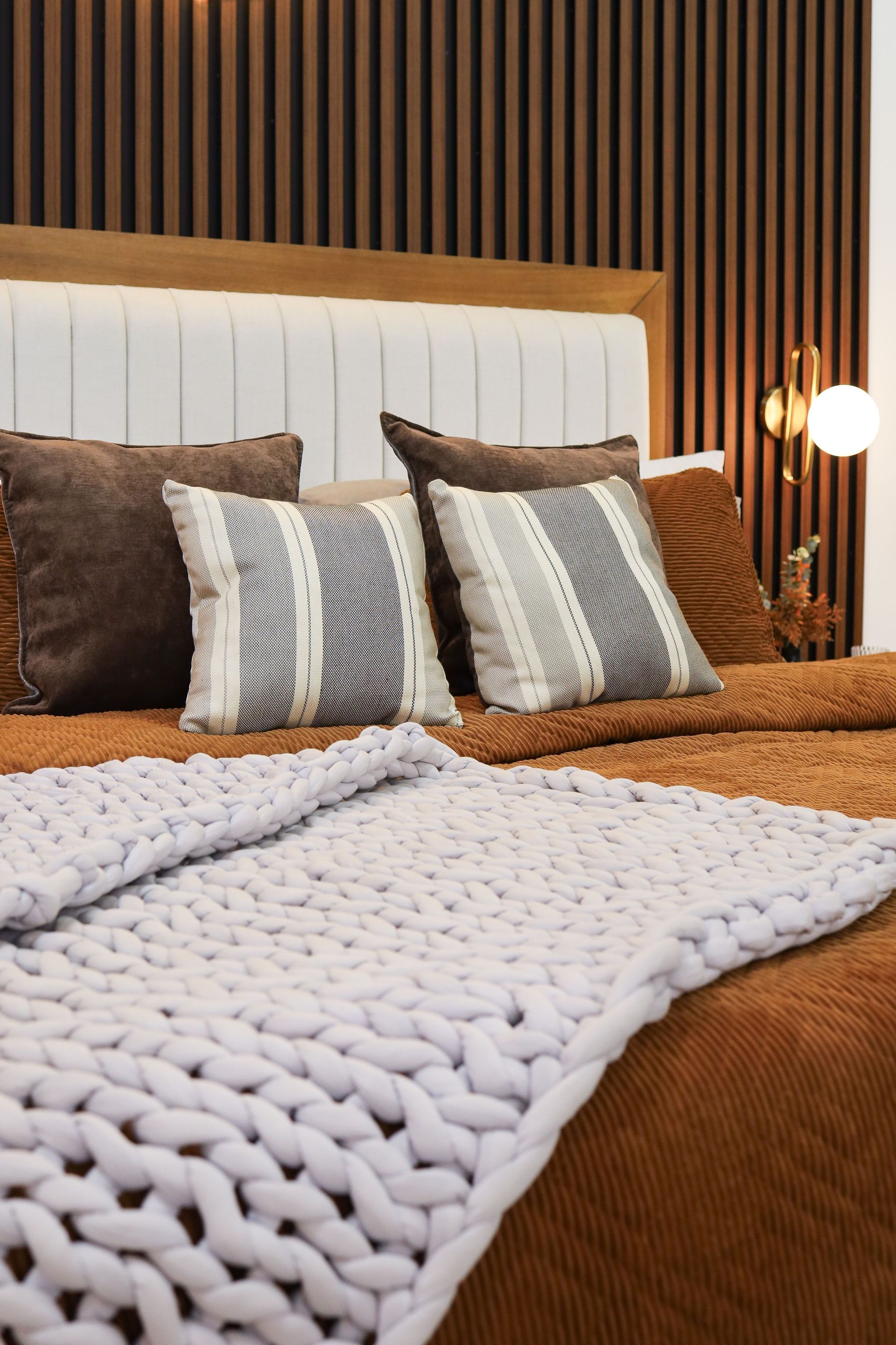 Cozy bed with brown and white pillows, a chunky knit throw, and a wood slat headboard lit by a warm lamp.