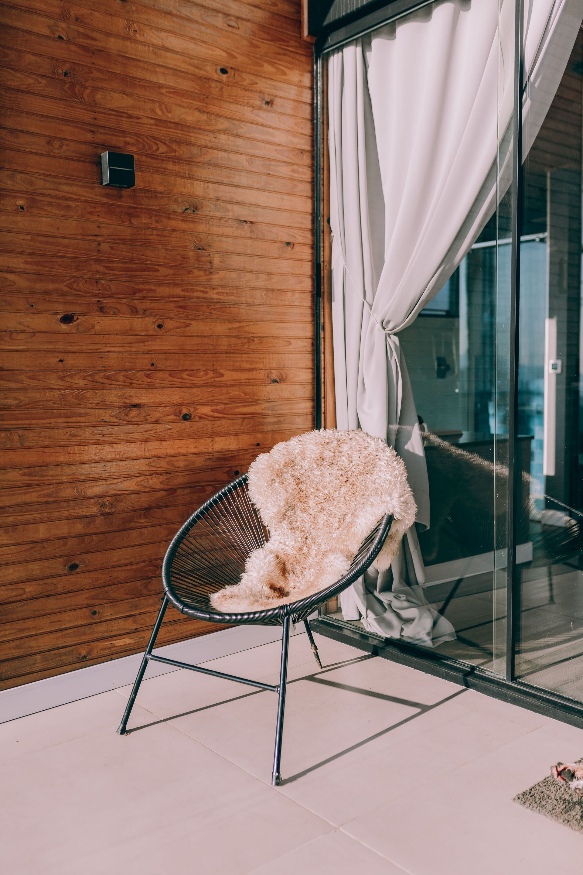 Black wire chair with fluffy white cushion on a tiled balcony beside a wooden wall and glass door.