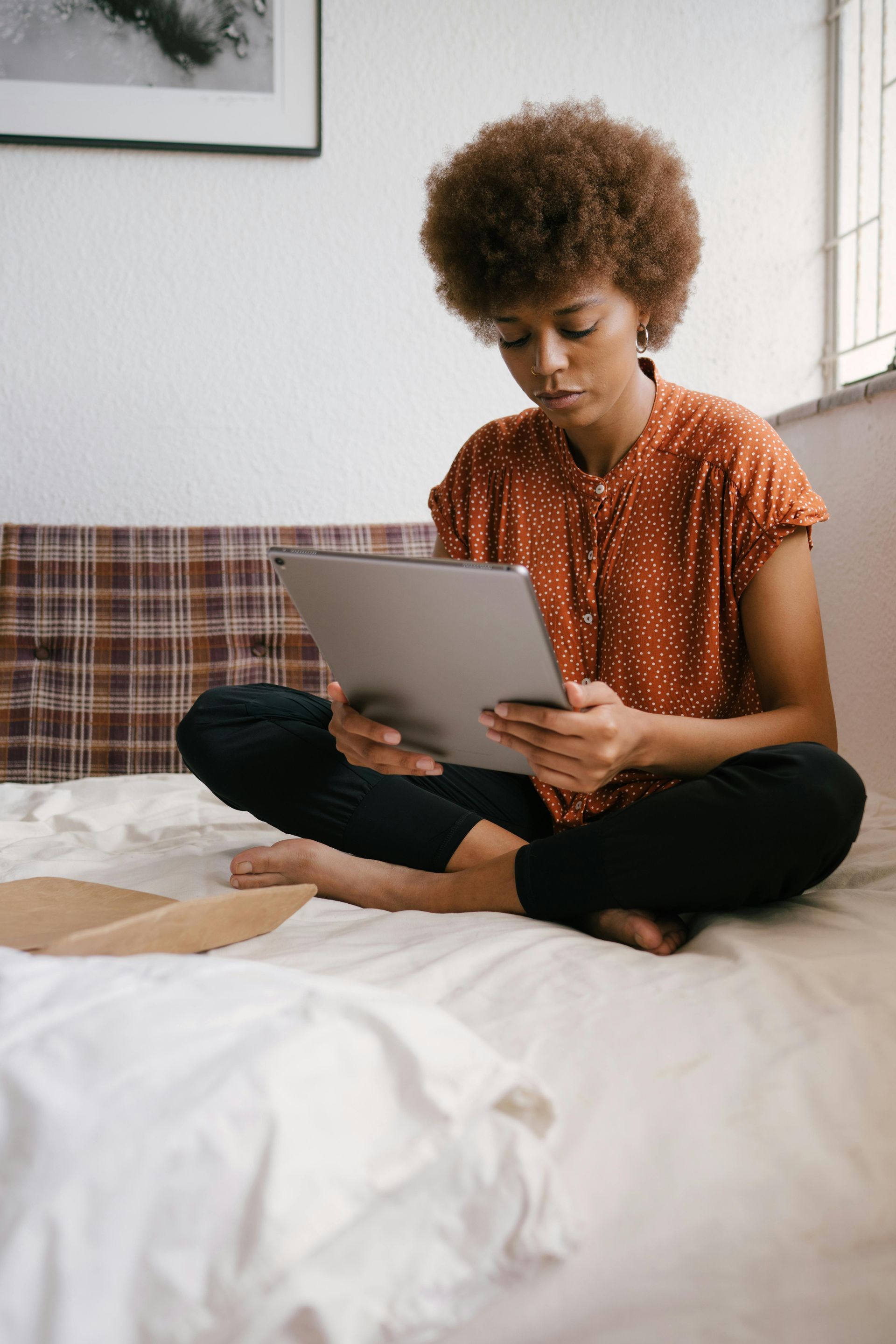 Woman with afro sits cross-legged on bed, looking at tablet, wearing orange shirt and black pants.