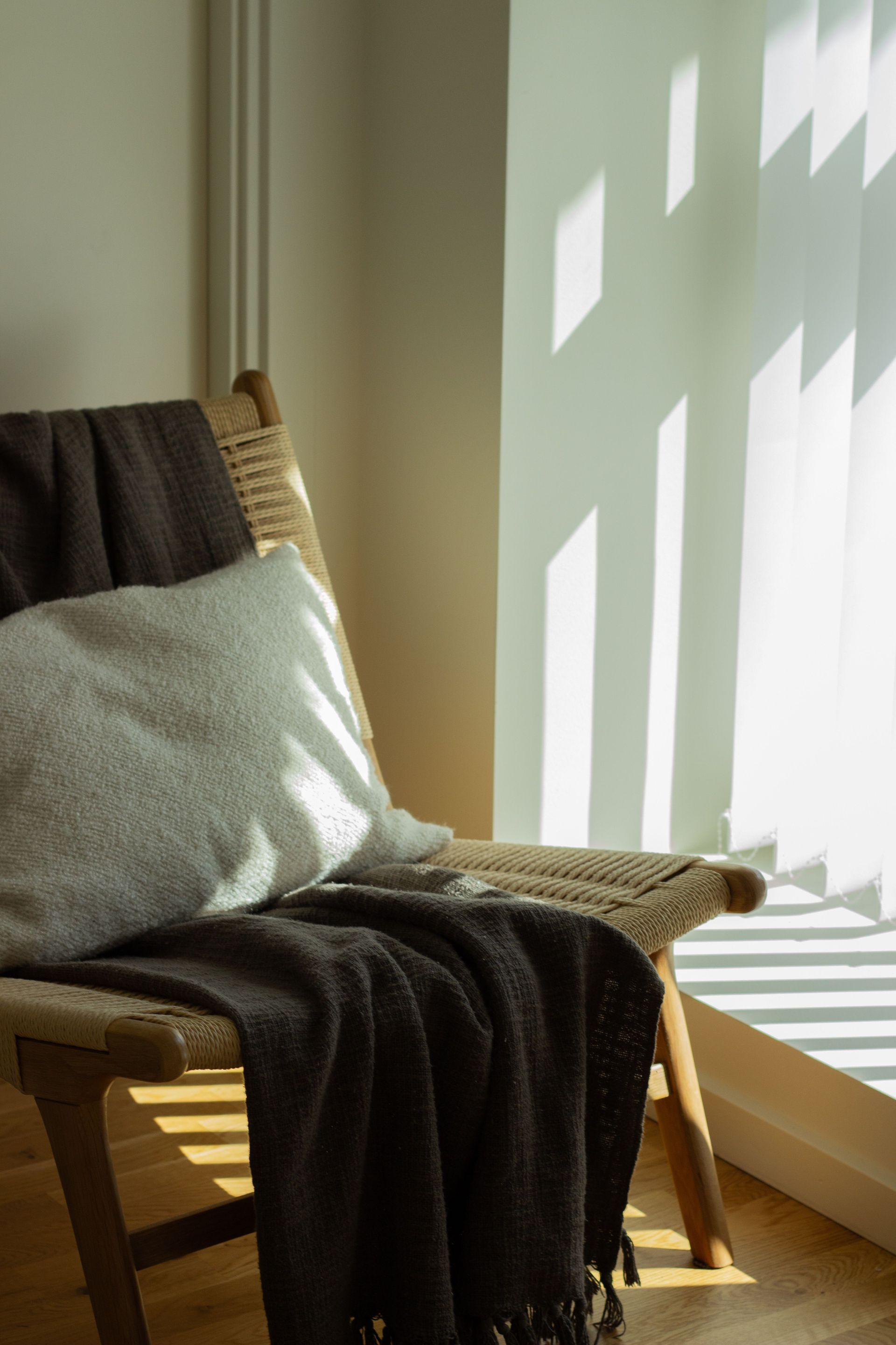 Wicker chair by a sunlit window with a gray pillow and dark blanket draped over it