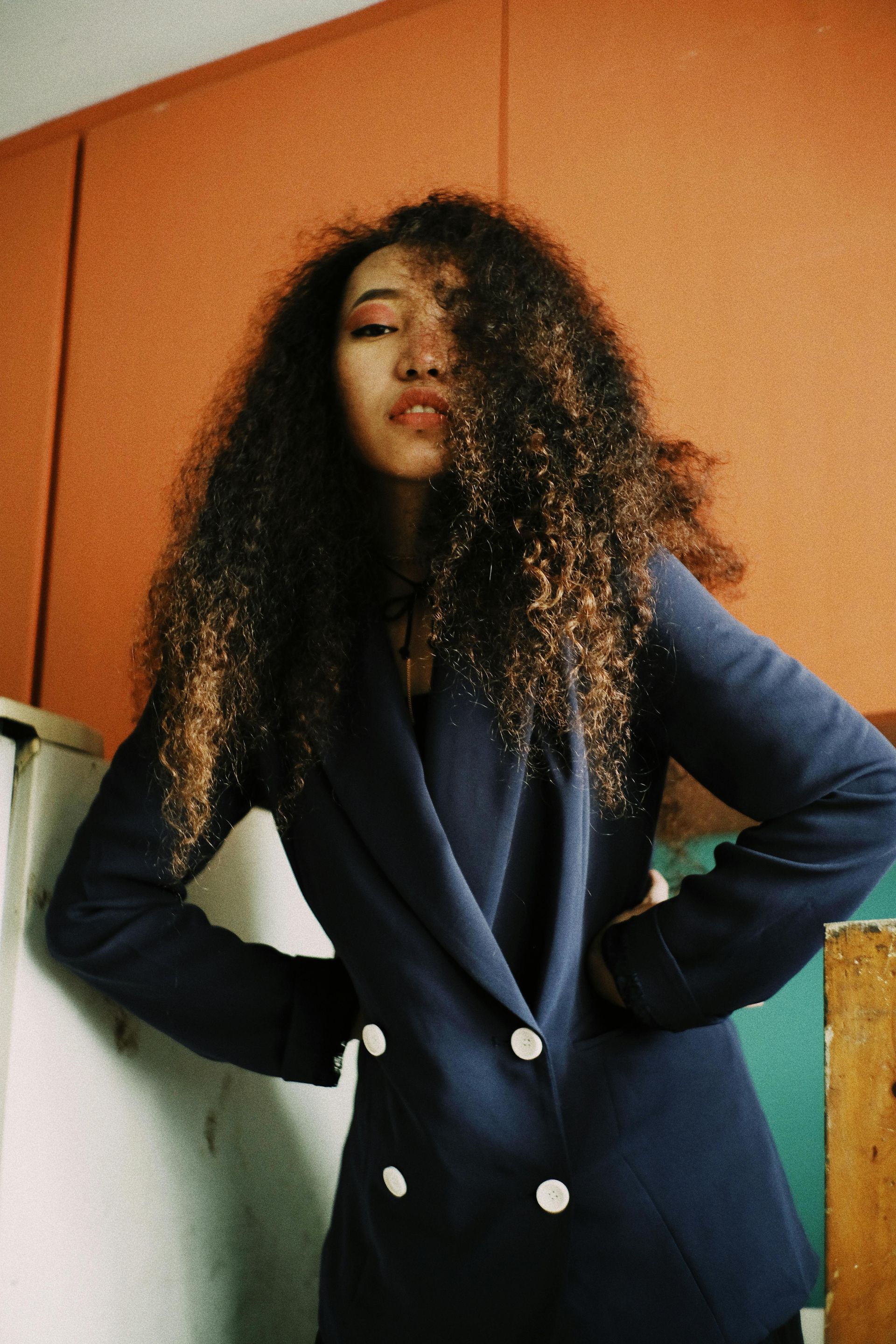 Woman with curly brown hair, in a navy blazer, hands on hips, orange background.