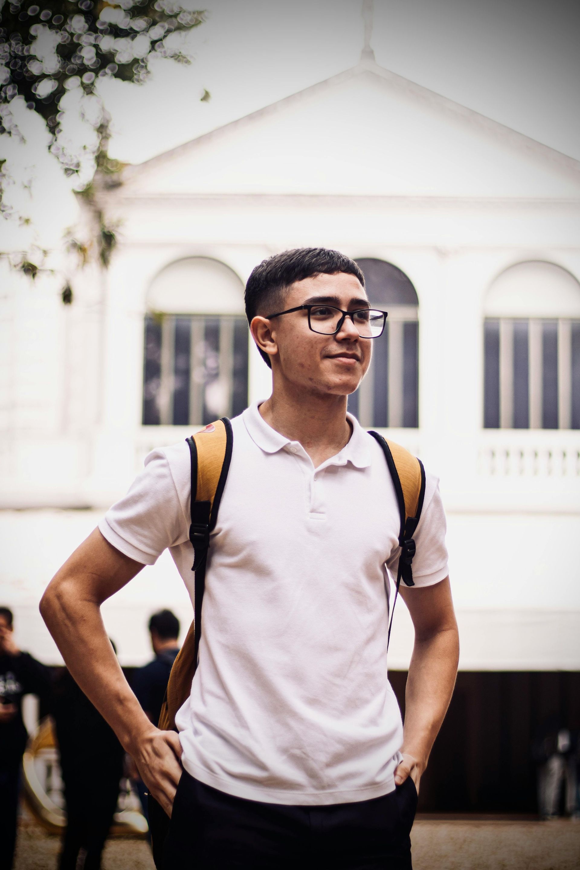 Young man with glasses, backpack, and white polo shirt, standing outside a white building, hands in pockets.