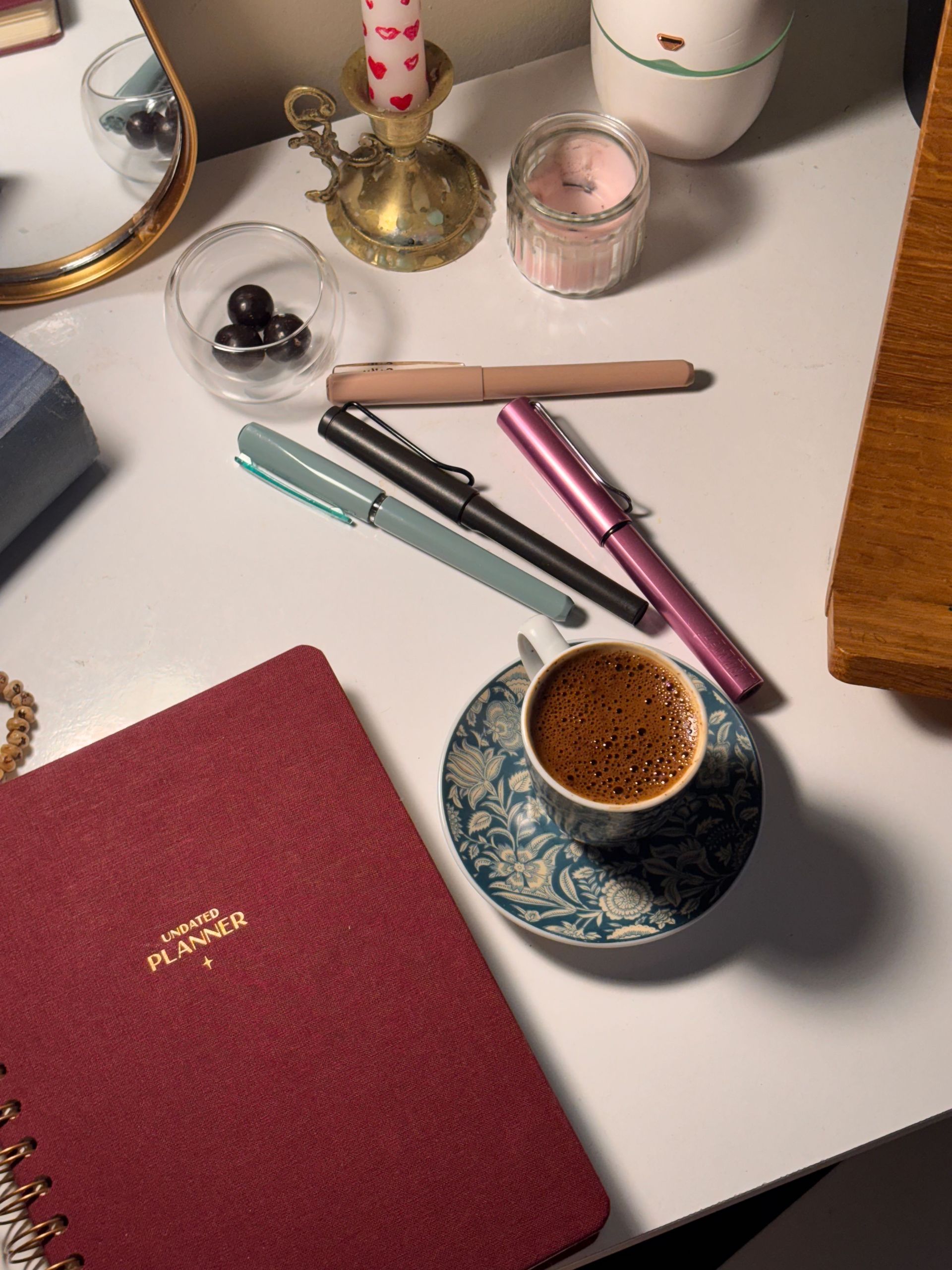 Desk with three pens, a coffee cup, red notebook, candles, and small decorative items on a white surface.