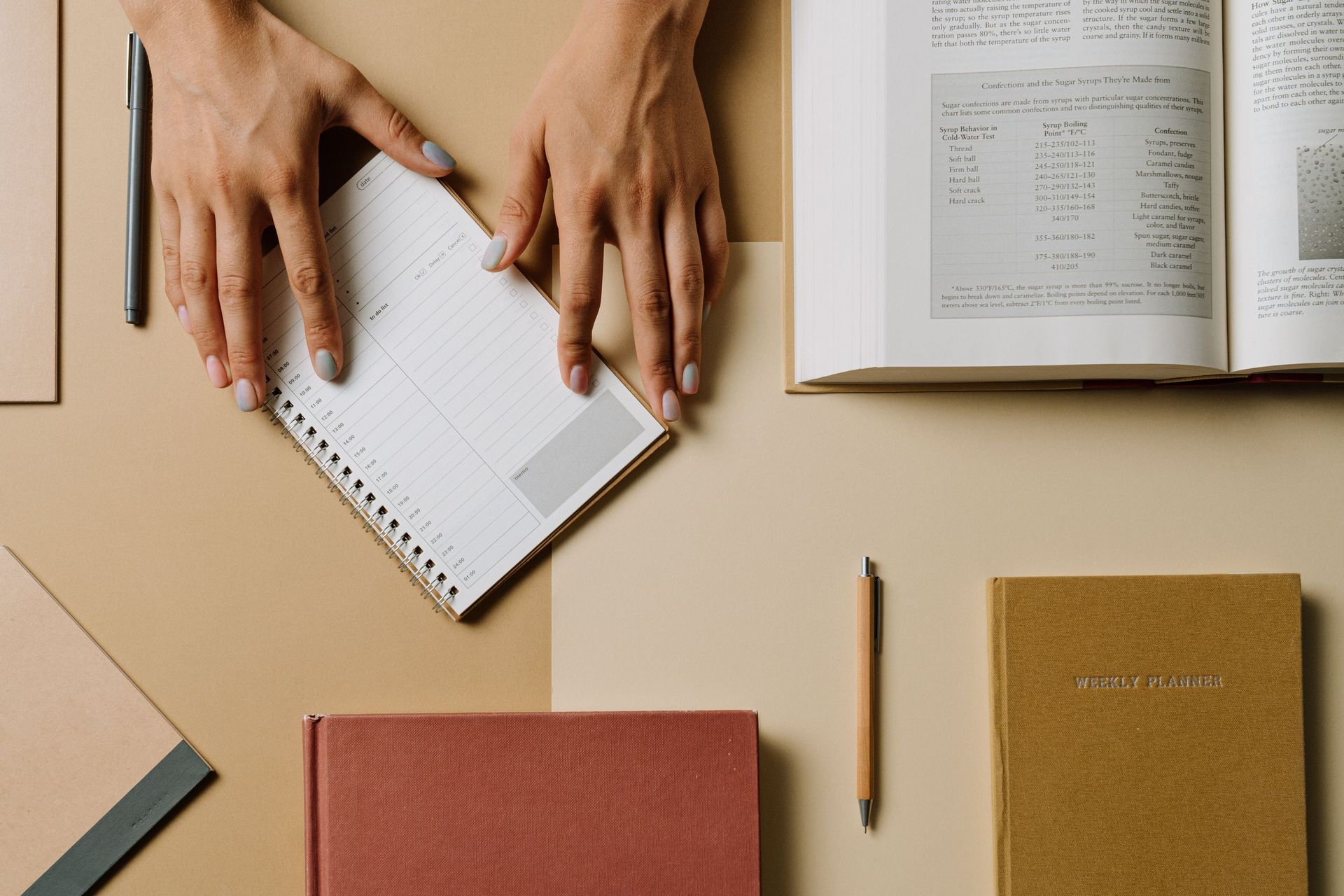 Hands writing in a spiral notebook on a beige desk with books, pencil, and notebooks around it