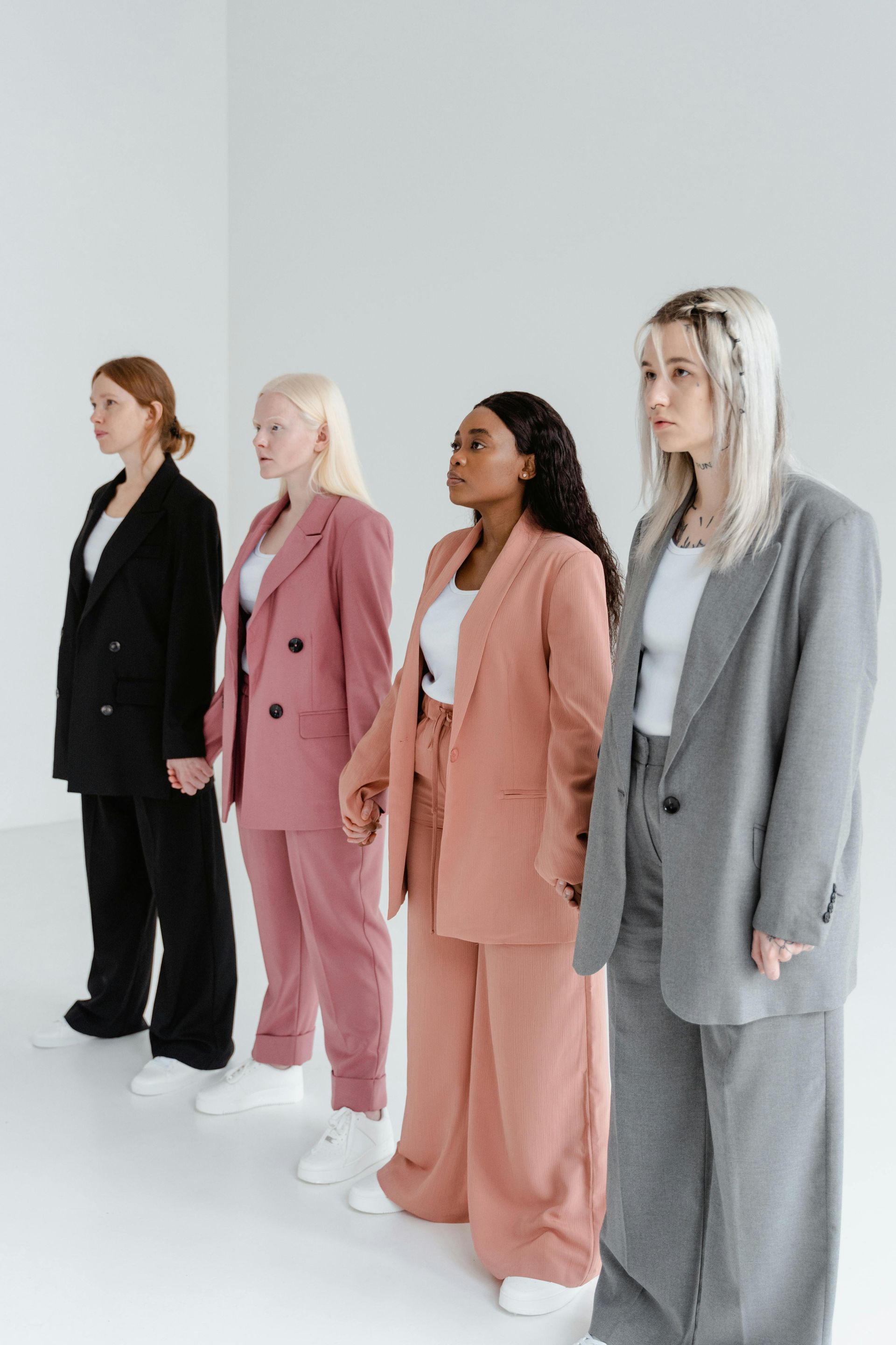 Four women in suits, various skin tones and hair colors, holding hands, posing in a white room.