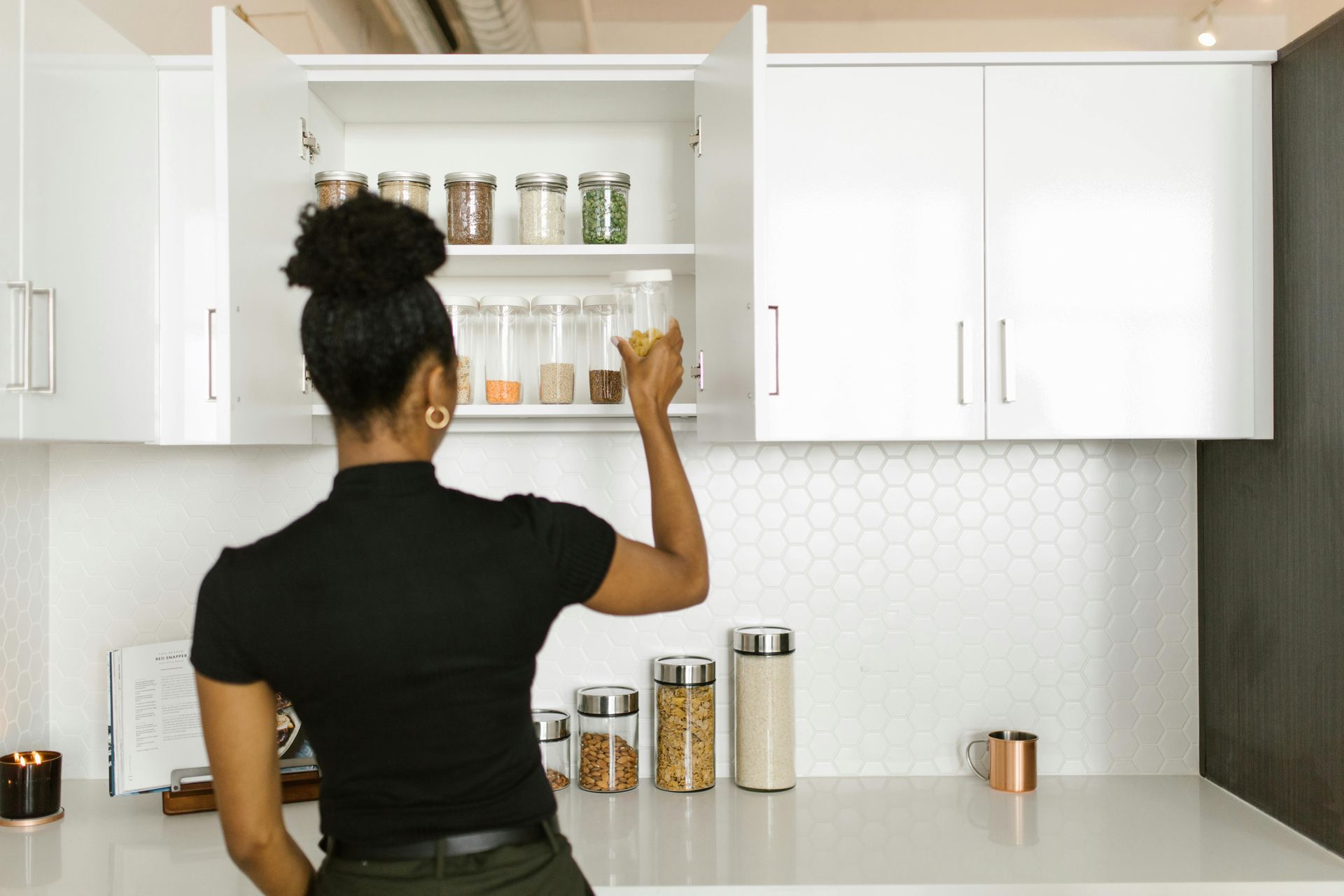 Woman reaching into an open kitchen cabinet for a jar; white cabinets, countertop, and backsplash.