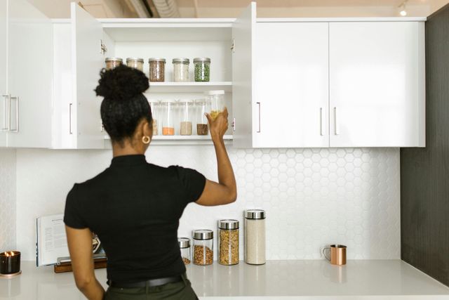 Woman reaching into an open kitchen cabinet for a jar; white cabinets, countertop, and backsplash.