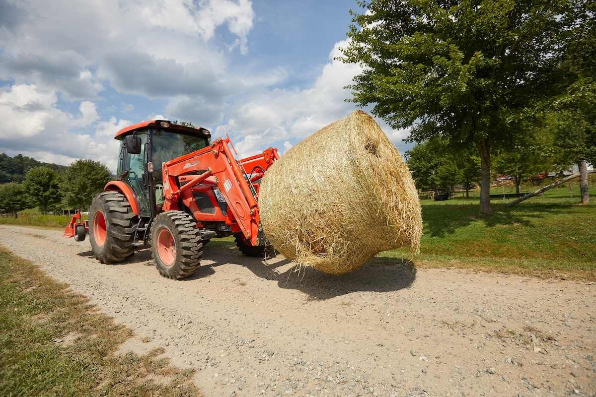 Red Tractor Carrying A Large Round Hay Bale On A Gravel Farm Road — Thompson's Farm Gear In Wauchope, NSW