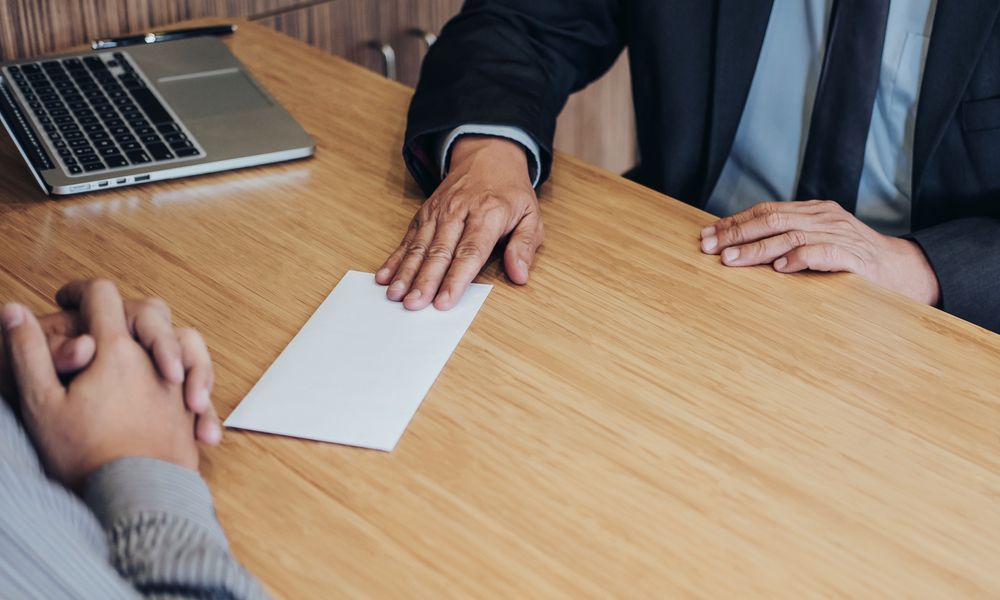 A person in a suit handing a white envelope to someone across a wooden table.