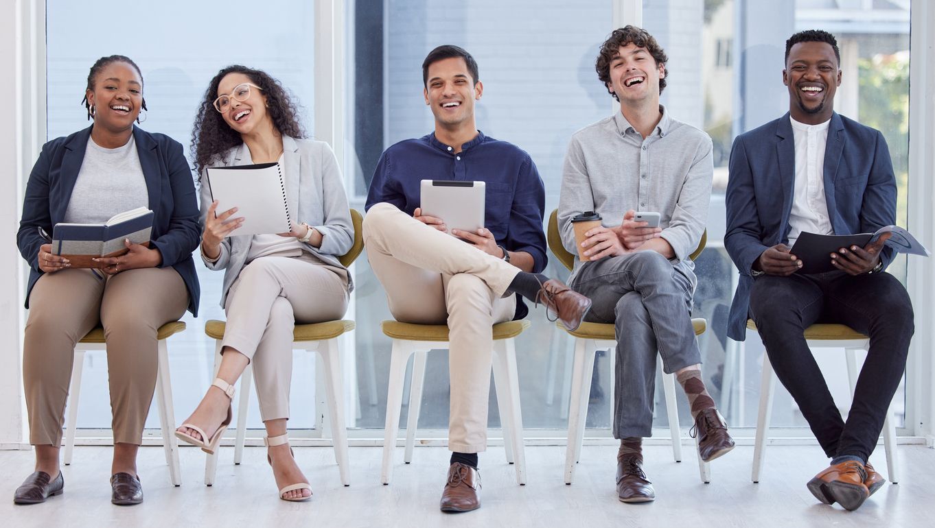 Five people in business attire sit in a row on chairs, holding tablets and folders while smiling in a bright office.