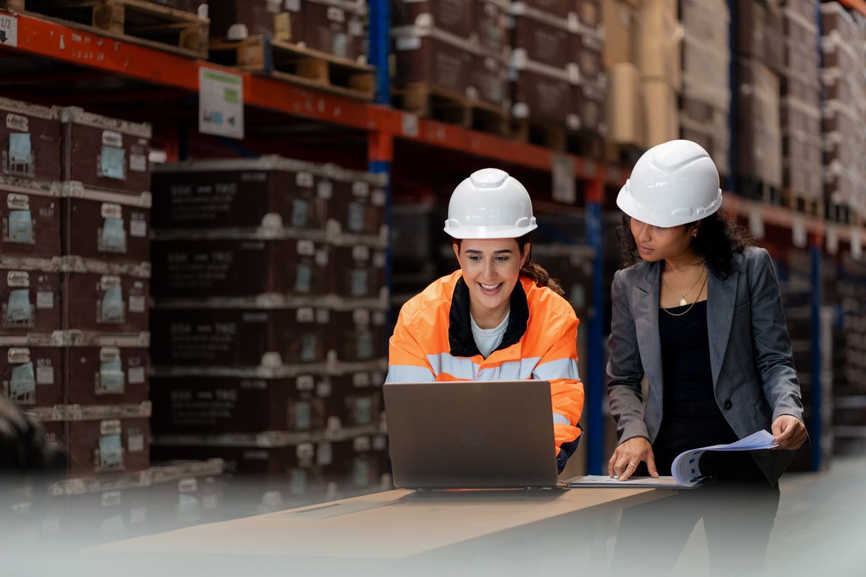 Two women in hard hats, one in orange vest, reviewing laptop and papers in a warehouse with stacked boxes.