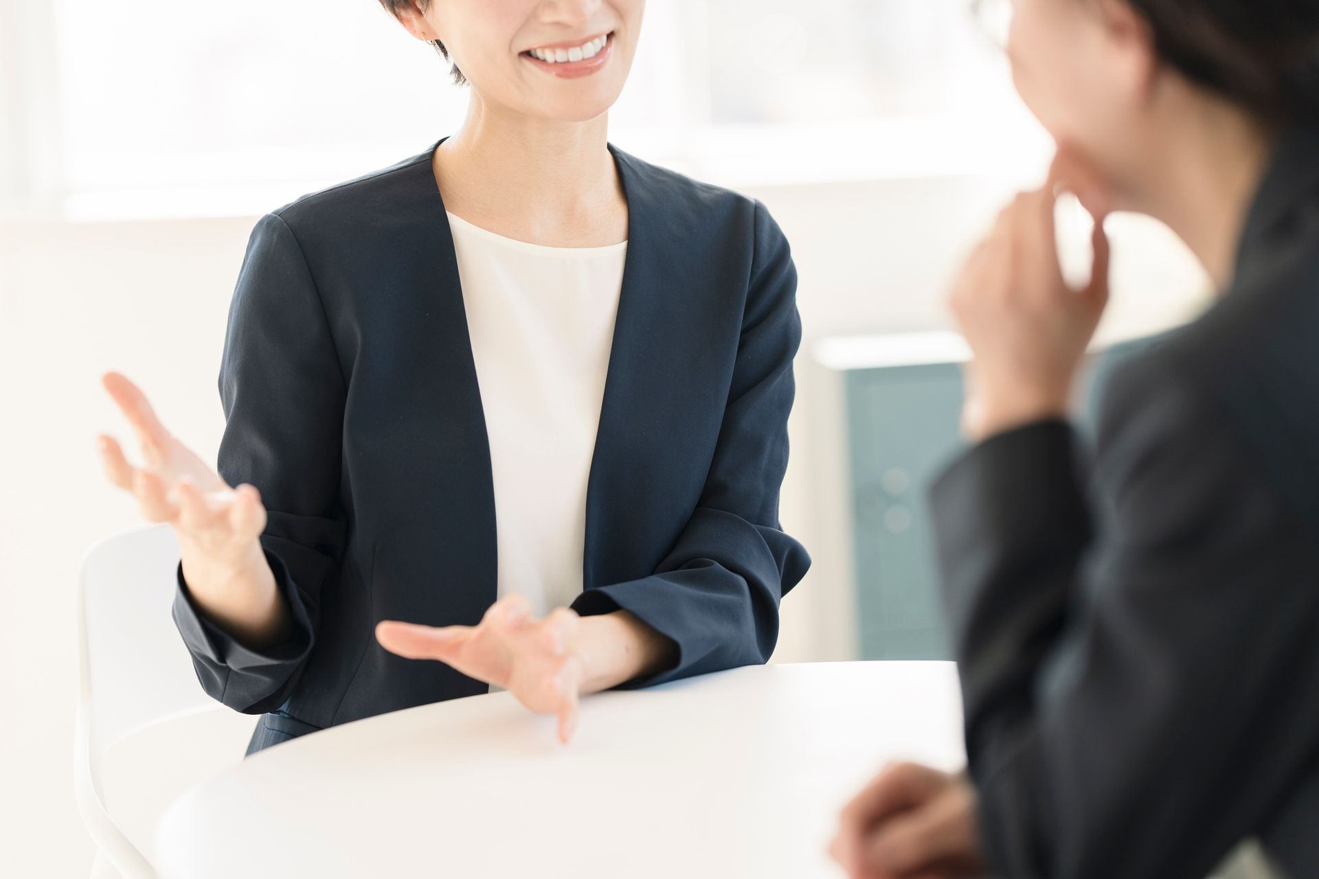 Two women in business attire seated at a table, one gesturing and smiling, the other listening.