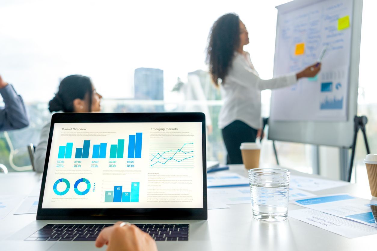 A professional meeting where a person presents at a whiteboard while a laptop in the foreground displays data charts.