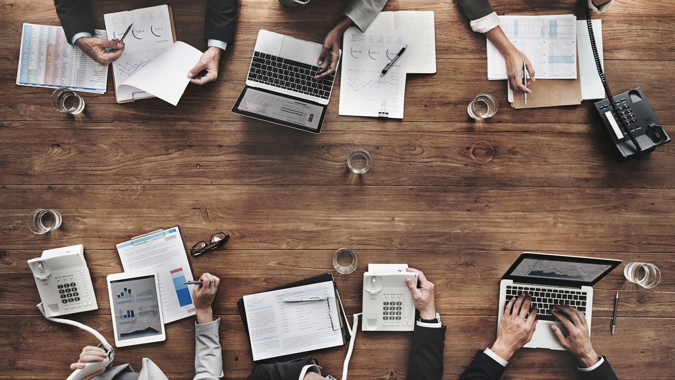 Business meeting at a wooden table, papers, laptops, phones, and hands in action.