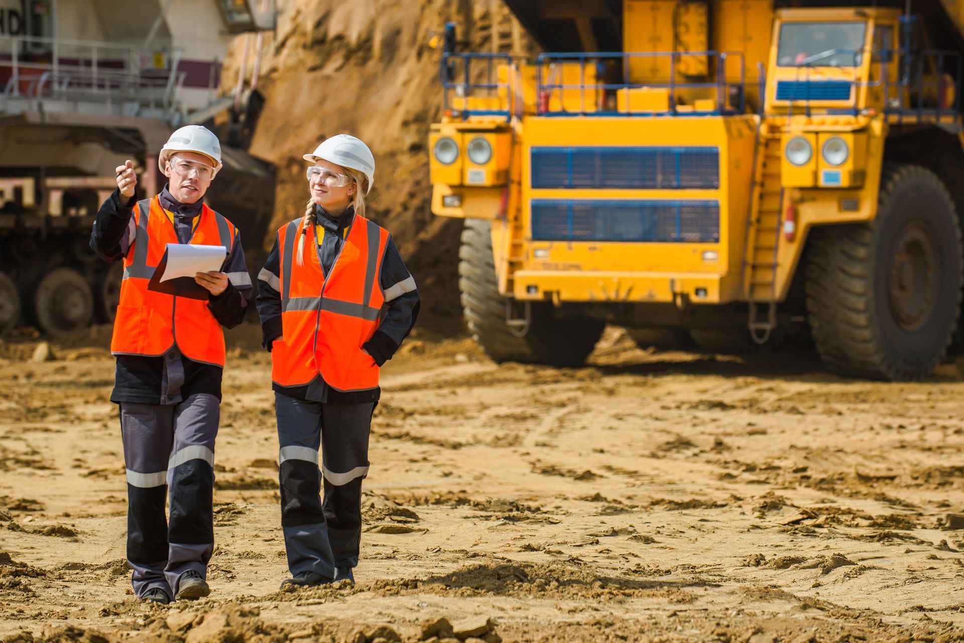 Two construction workers in orange safety vests stand in front of a large yellow dump truck.