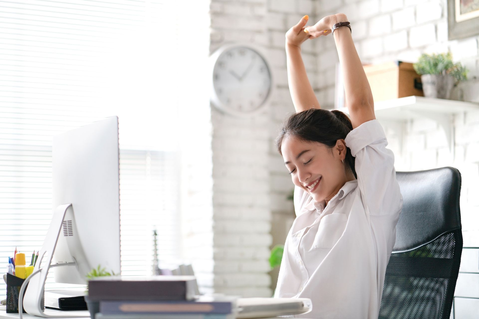 Woman stretching at her desk, arms raised, smiling. Office setting with computer and clock.