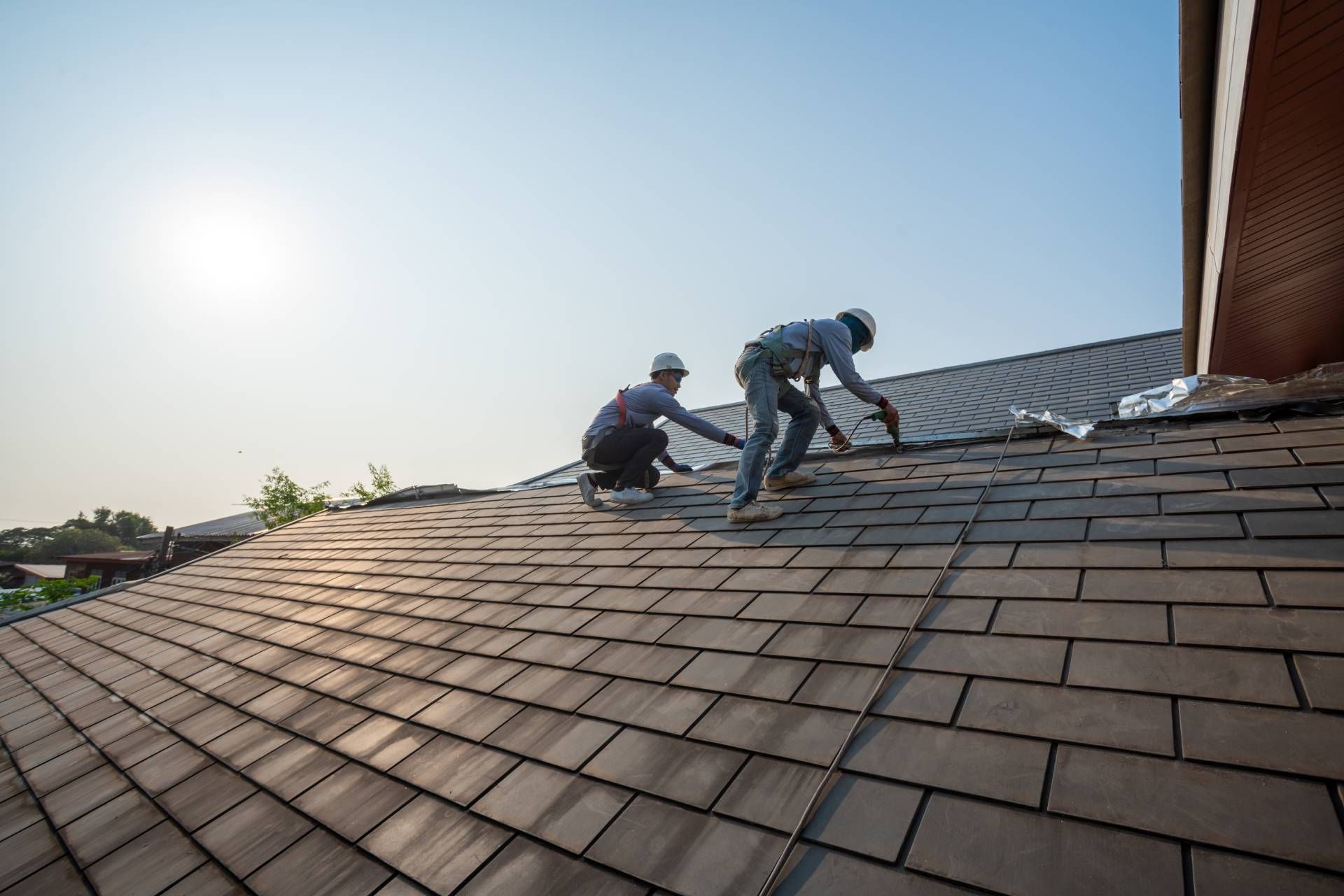 Two workers repairing roof shingles on a sloped rooftop in bright sunlight