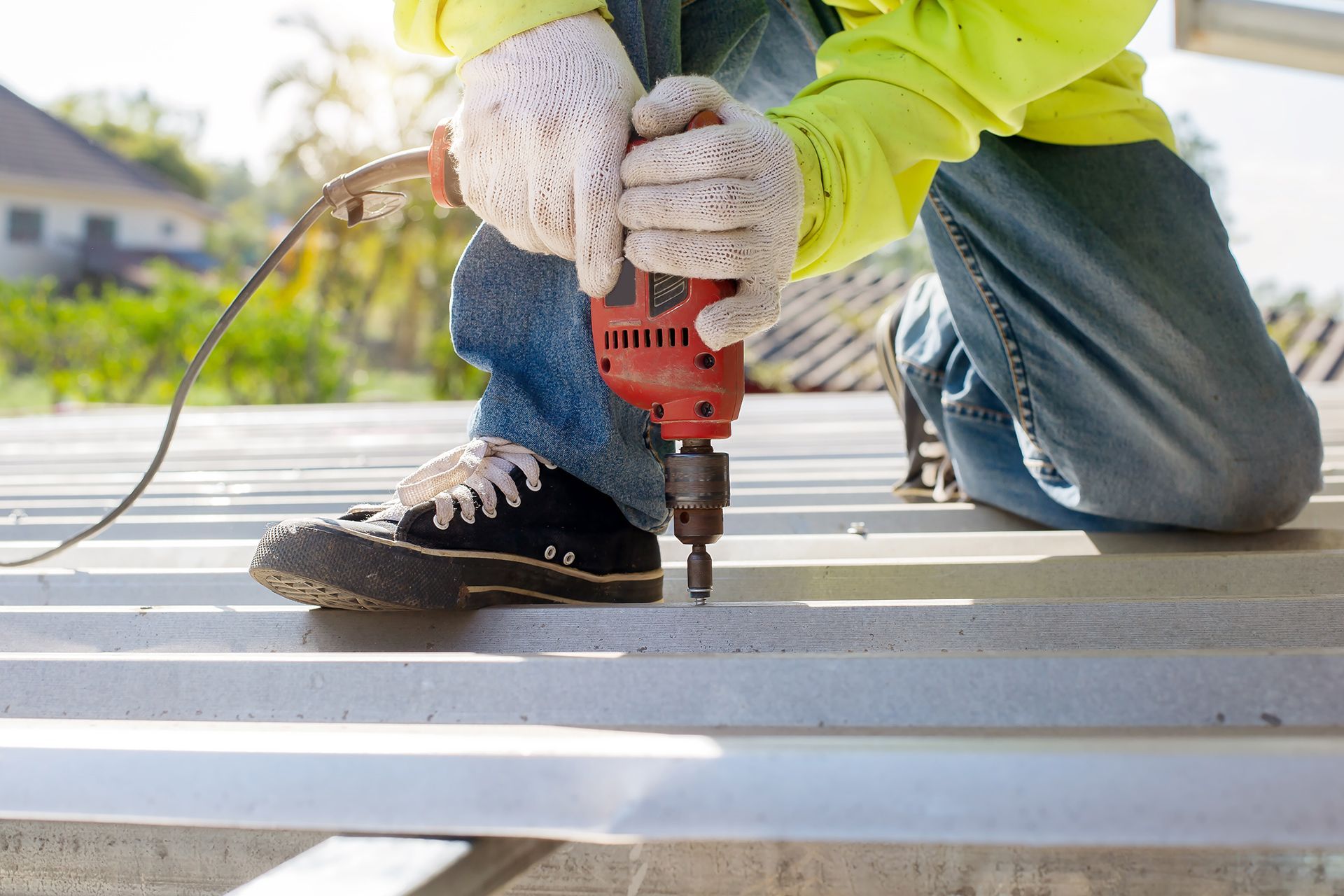 Worker drilling into a white metal surface outdoors, wearing gloves and jeans.