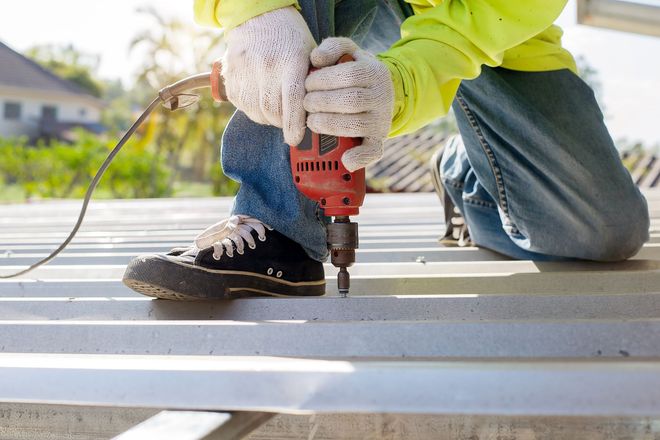 Worker drilling into a metal beam outdoors while kneeling in gloves and jeans.