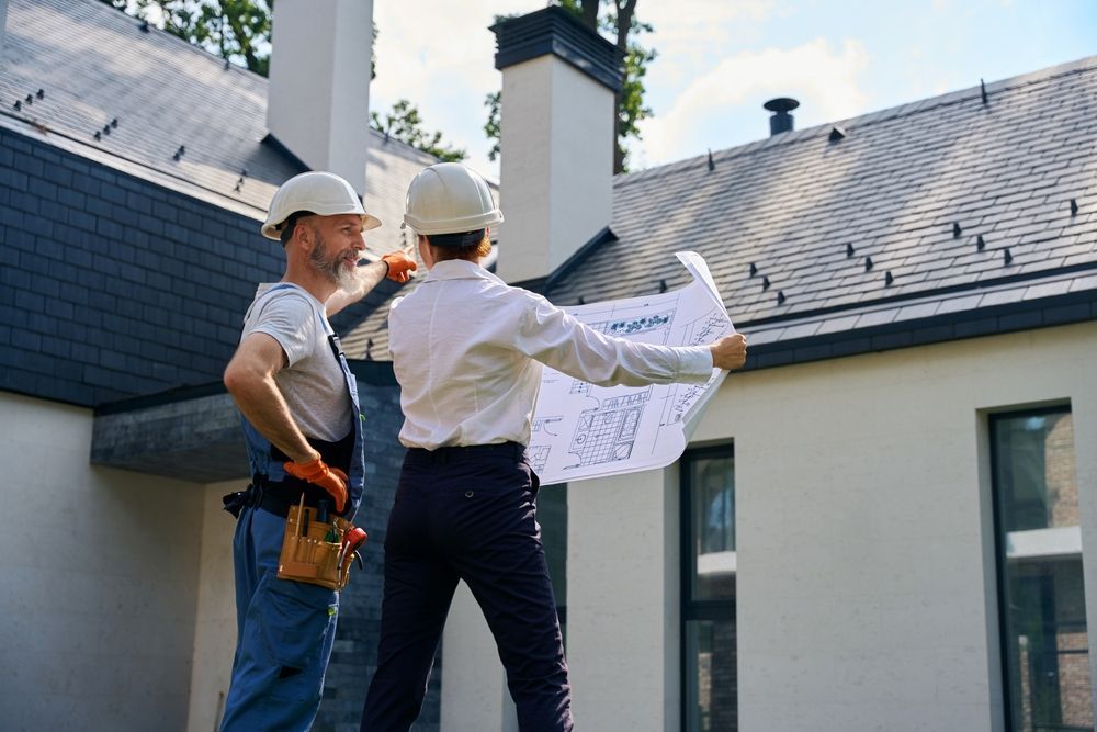 Two builders in hard hats review house plans beside a modern home exterior.