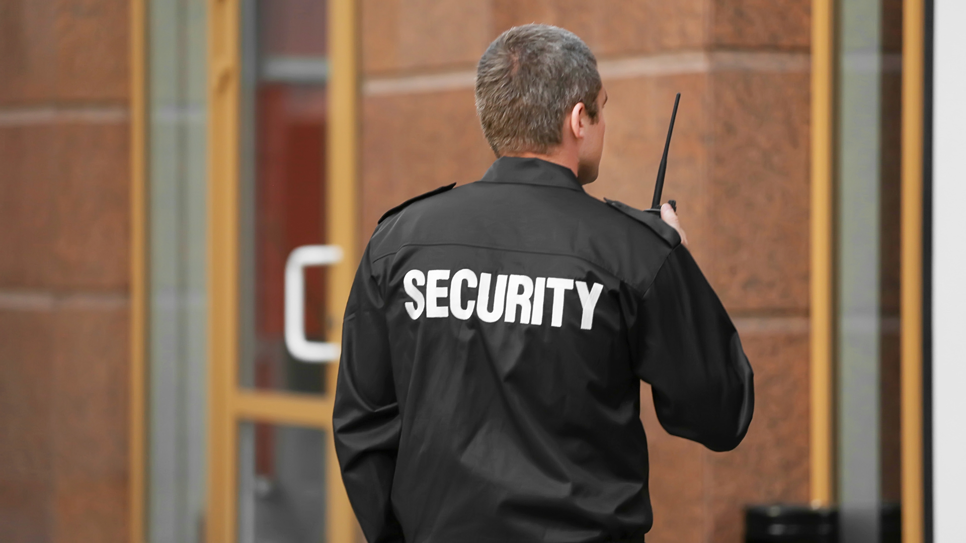 A security guard is standing in front of a building with a walkie talkie in his hand.