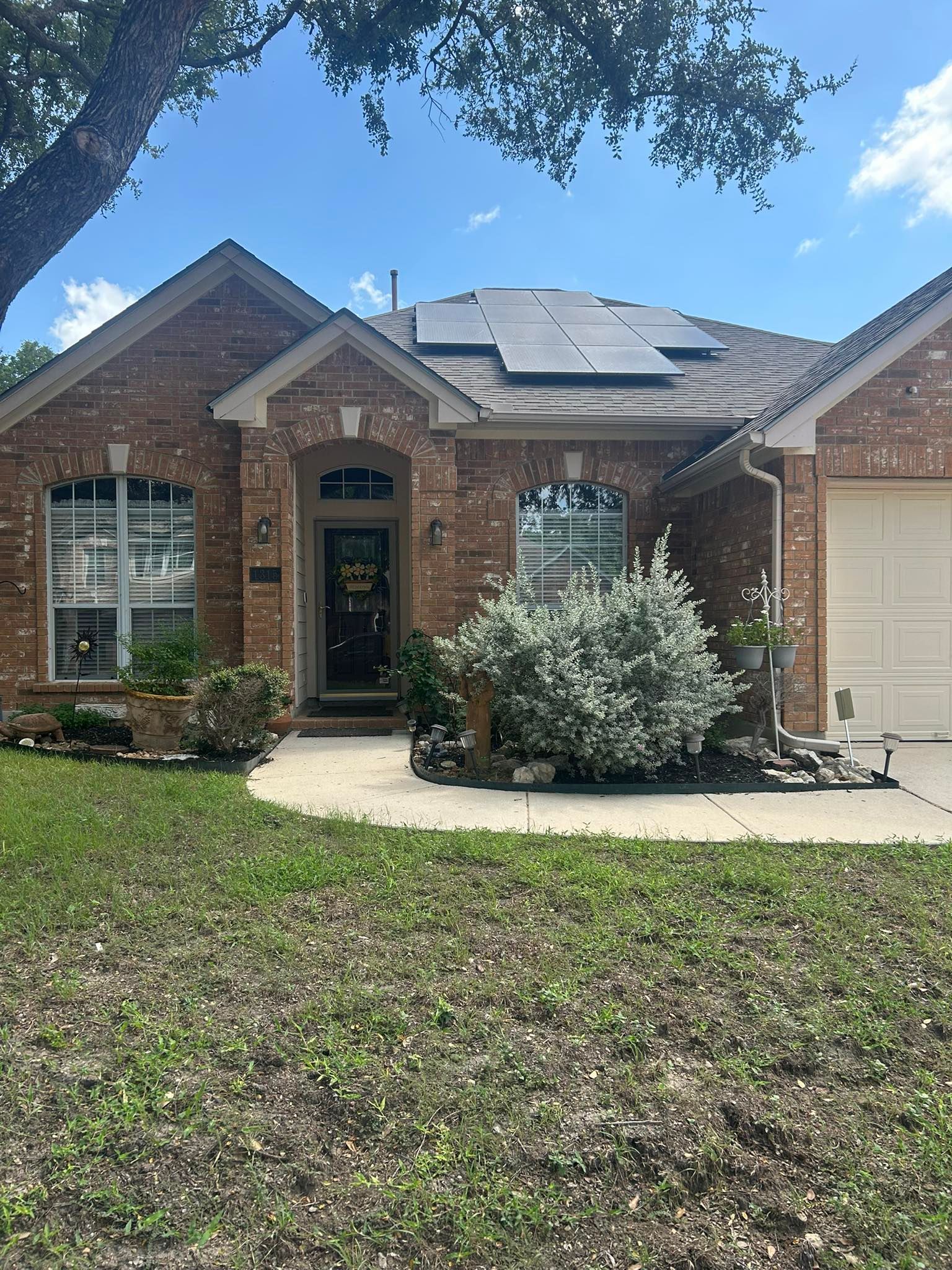 A single-story brick house with a front porch, a bush in the yard, and solar panels on the roof under a blue sky.