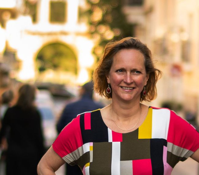 De schrijfdocent staat in een kleurrijk shirt en lacht naar de camera