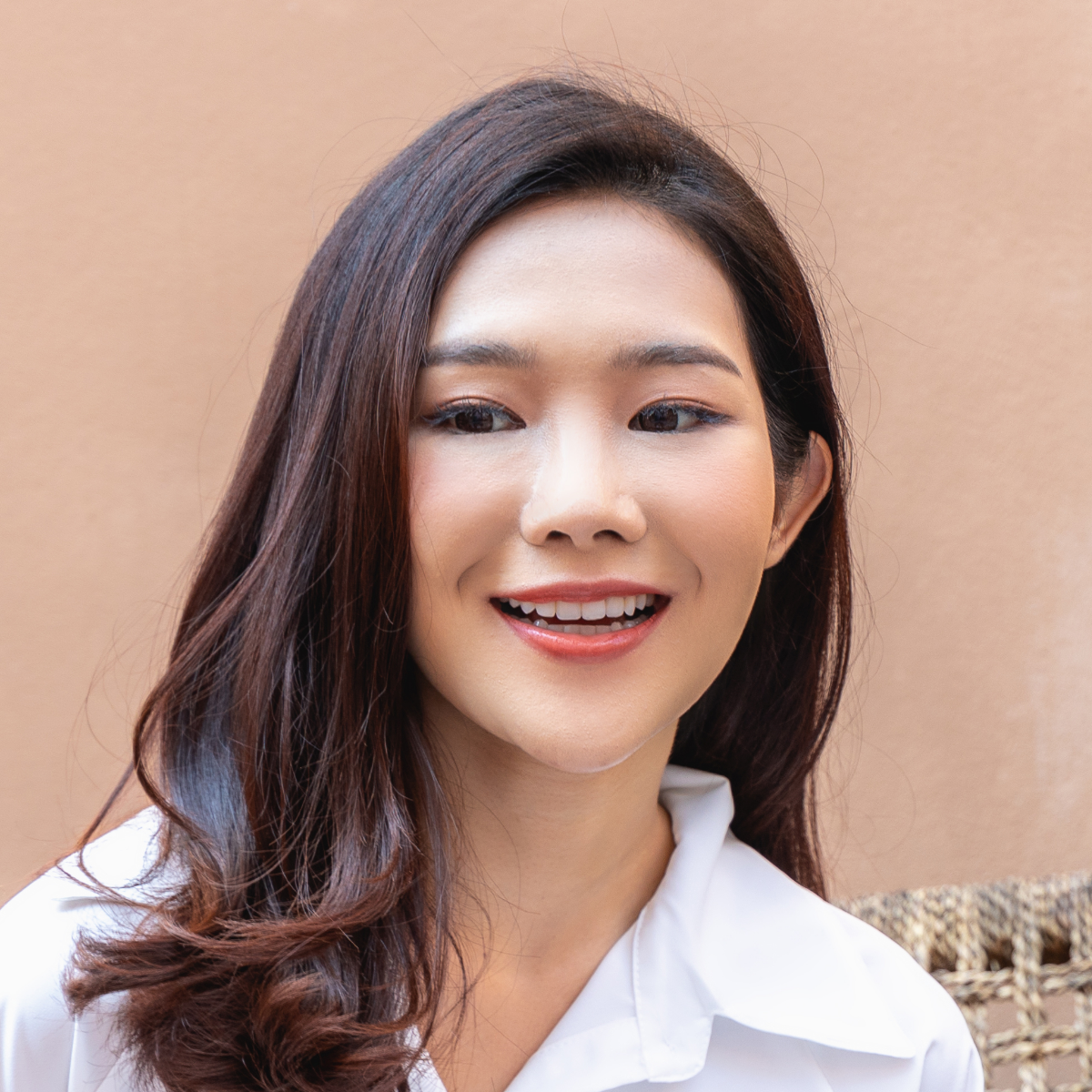 Woman with dark hair smiles, wearing a white shirt; outdoors against a tan wall.