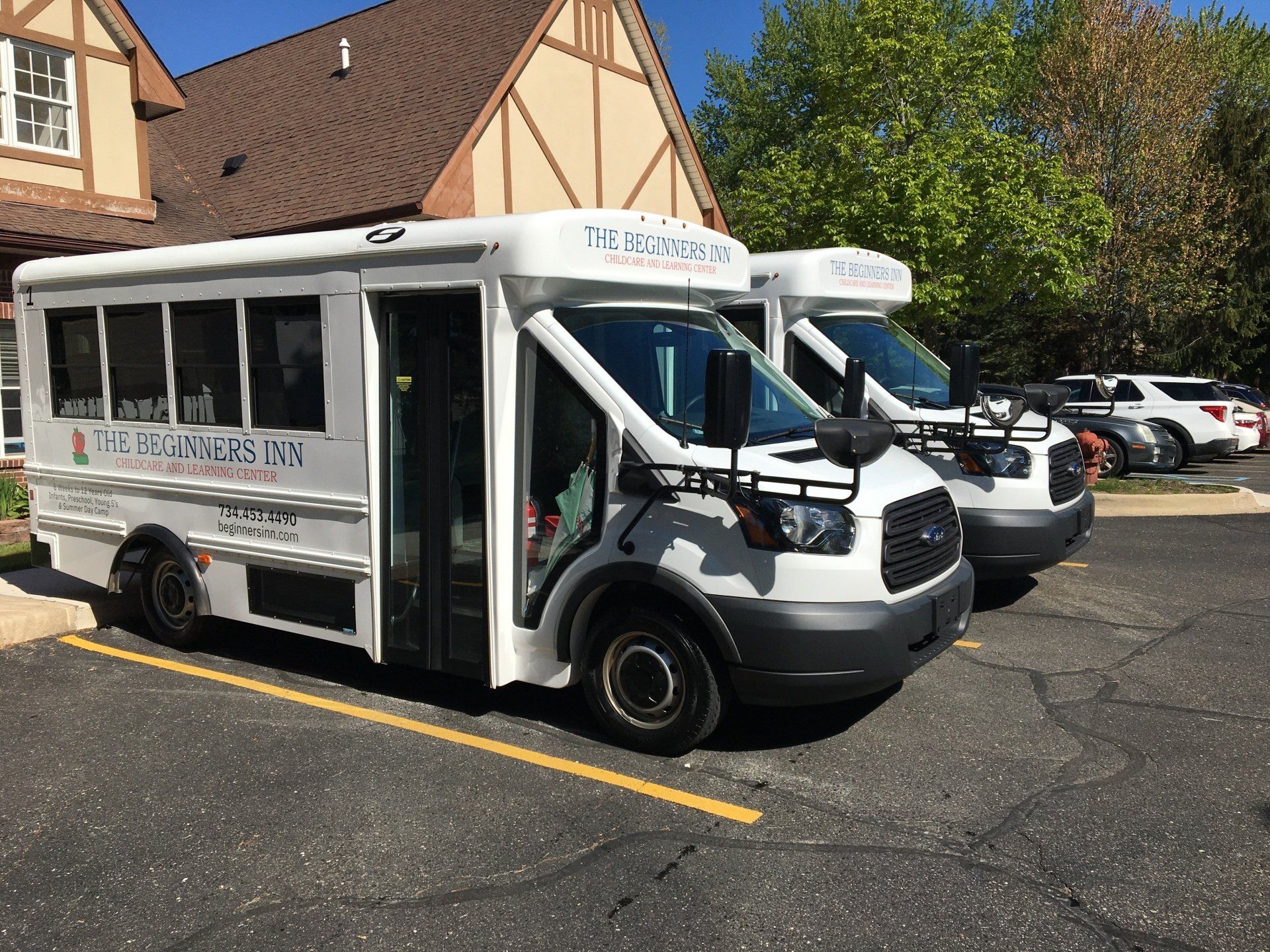 Two white passenger buses parked in front of a building, possibly for a school or daycare. Sunny day.