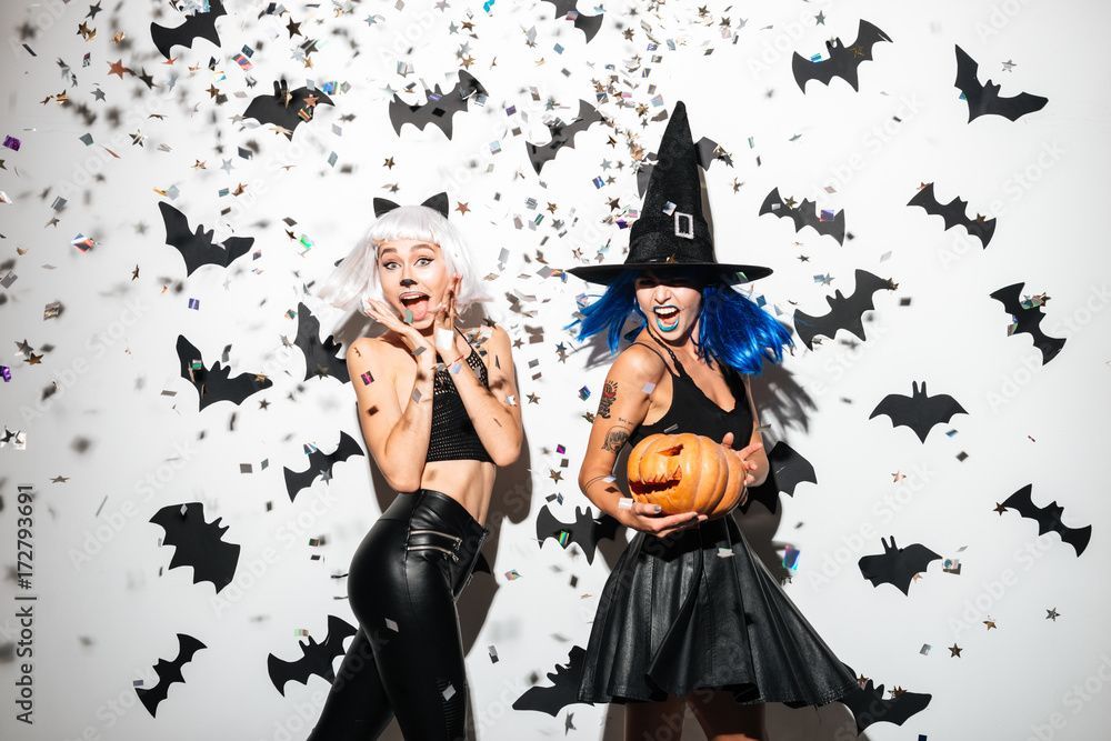 Two women in Halloween costumes surrounded by bats and confetti.