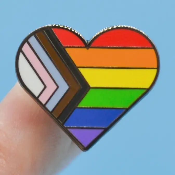 Heart-shaped enamel pin with rainbow Pride stripes and pink, white, blue, and brown chevrons on a finger