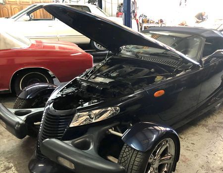 A blue Plymouth Prowler with its hood open in a garage. A red car is visible in the background.