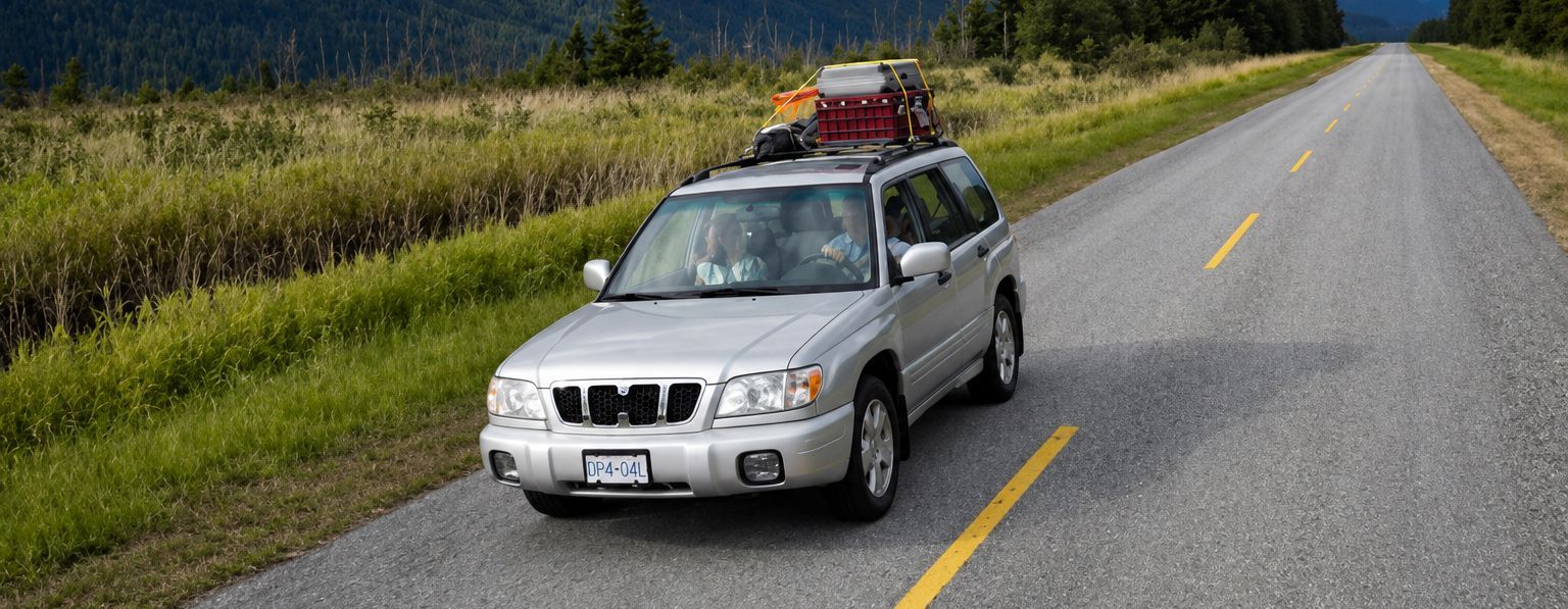 Silver SUV with roof cargo driving on a rural road beside grassy fields and forest.