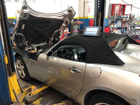 A silver convertible car in a garage with the hood open. A diagnostic computer sits on the roof.