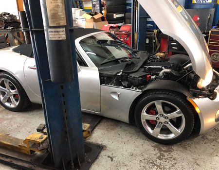 Silver convertible car with its hood open in a mechanic's shop, revealing the engine.