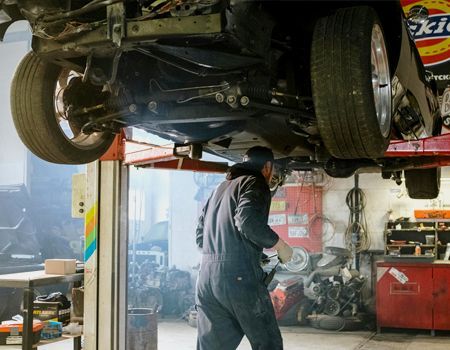 Mechanic working under a car on a lift in a garage; interior setting, work overalls.
