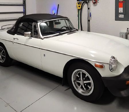 White MG MGB convertible with a black top parked in a garage. The car has chrome trim and black wheels.