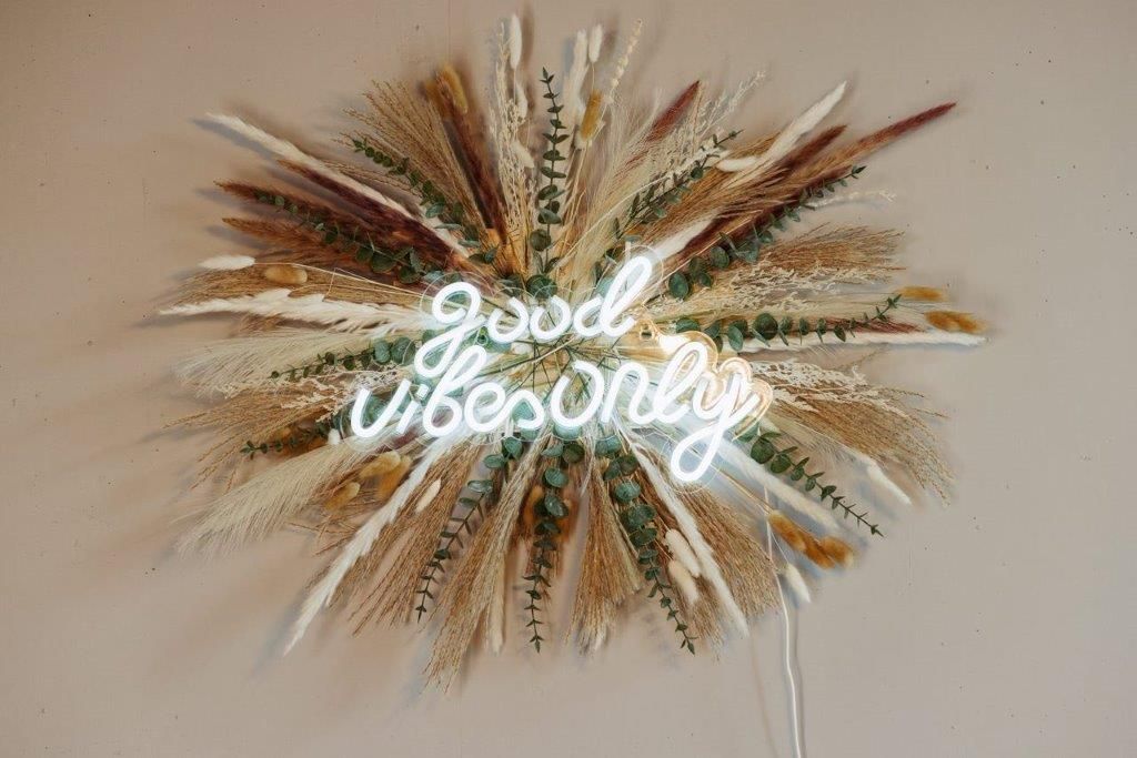 Ein Neonschild mit der Aufschrift „Nur gute Stimmung“ ist von getrockneten Blumen an einer Wand umgeben.