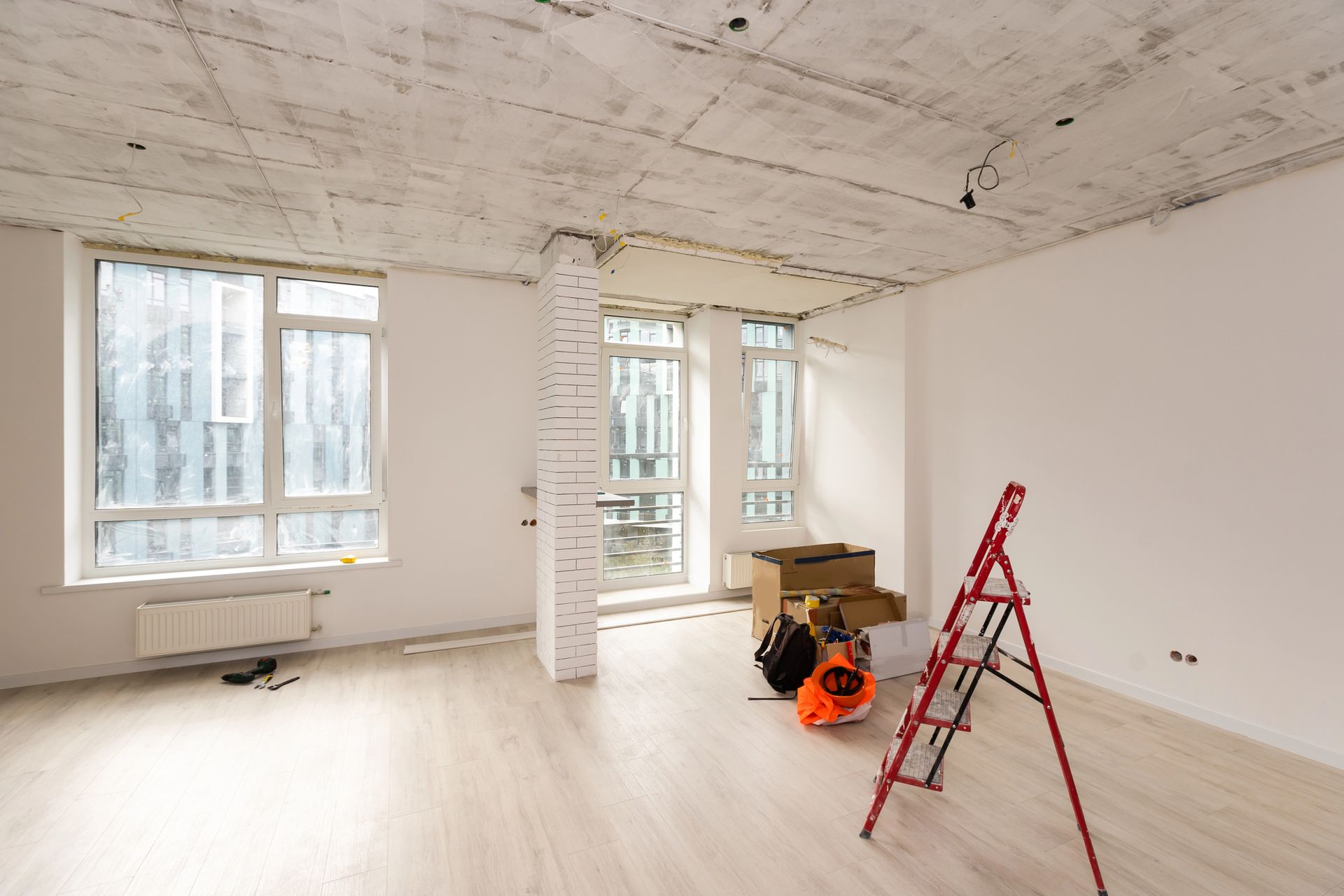 Una stanza vuota in fase di ristrutturazione presenta un soffitto in cemento, pareti bianche, ampie finestre e una scala a pioli rossa.