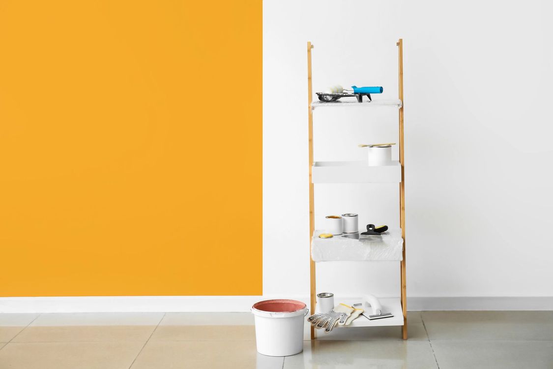 Una parete parzialmente dipinta di giallo si erge accanto a una scaffalatura in legno che contiene barattoli di vernice e altri materiali, su un pavimento piastrellato.