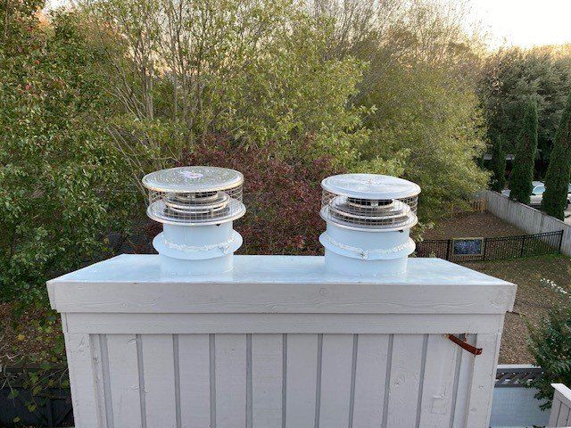 Two white chimney vents with wire cages on a white rooftop against a background of trees and a fence.