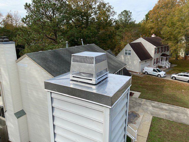 Close-up of a chimney with a silver metal cap on a residential roof. A car and trees are in the background.