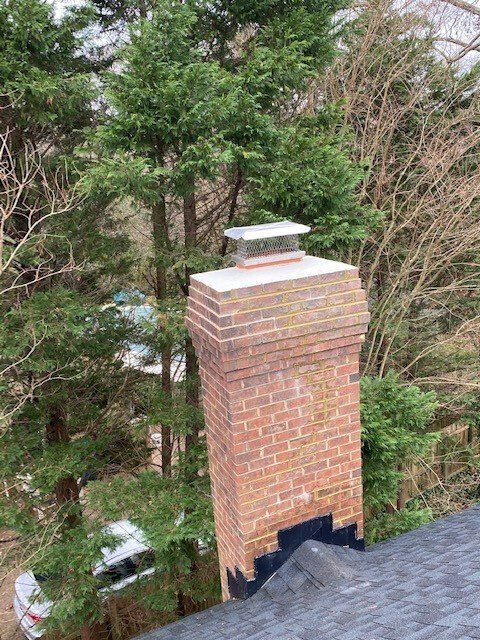 A brick chimney with a metal cap, set against a backdrop of trees. The roof below is dark gray.